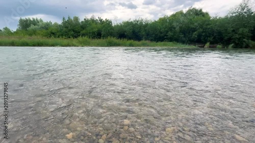 A calm river flows past green grassy banks under a cloudy sky. Dense summer trees grow along the opposite shoreline in daylight. The water surface ripples gently through the scenic landscape.