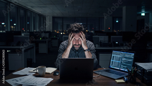 Stressed businessman sitting alone at his desk in a dark office late at night with laptops and papers scattered around him looking frustrated and overwhelmed.