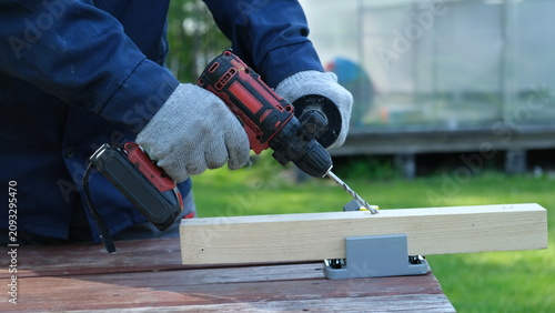 Photos Close-up of a worker in a blue jacket and protective gloves using a cordless electric drill to make a hole in a wooden beam outdoor