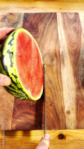 Vertical video social media - Closeup of a man’s hands using a large knife to cut a whole juicy water melon or watermelon in half on a chopping board, with a thick green skin and pink fruit inside.