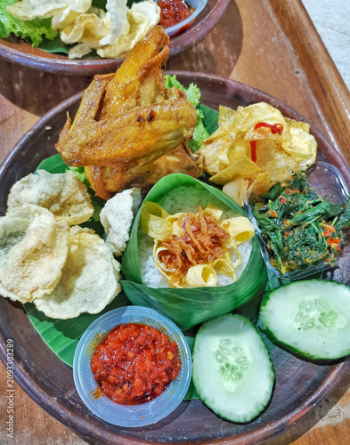 Authentic Indonesian fried chicken served with steamed rice wrapped in banana leaf, sambal, vegetables, cucumber and crackers. Traditional Southeast Asian comfort food on rustic wooden table.