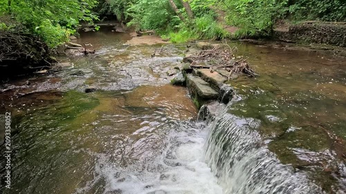 Water cascades down a woodland waterfall into a gentle stream as the camera slowly pans across the scene in slow motion, revealing the tranquil forest and flowing water.