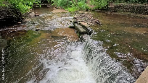 Water cascades down a woodland waterfall into a gentle stream as the camera slowly pushes in in slow motion, drawing focus to the peaceful flow and lush forest surroundings.
