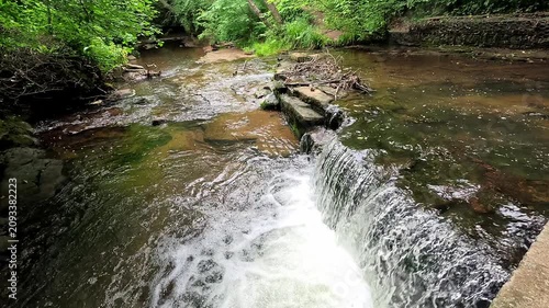 Water cascades over a small woodland waterfall into a gentle stream. Captured in a super slow-motion static shot, the scene emphasizes every ripple and splash amid the peaceful forest surroundings.