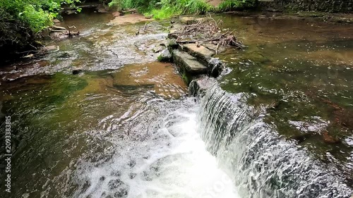 Water cascades over a small woodland waterfall into a gentle stream as the camera slowly pulls back in super slow motion, revealing the tranquil forest and serene natural surroundings.