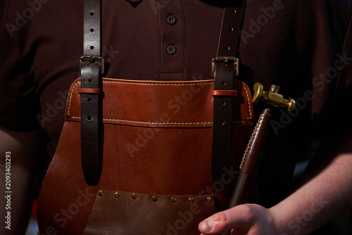 Obraz na plátně Leatherworker in a handcrafted brown apron holding a specialized brass hammer close up