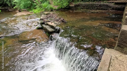 Stepping stones cross over a woodland waterfall as the camera slowly pulls back, revealing the flowing stream, mossy rocks, and peaceful forest surroundings in a tranquil natural setting.