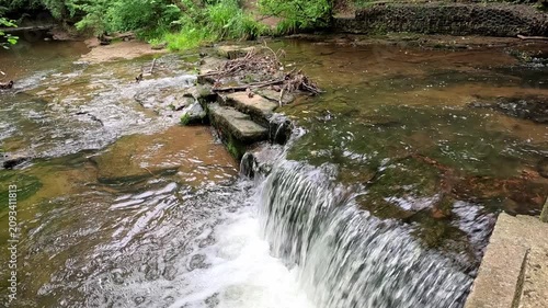 A panning shot reveals stepping stones crossing a woodland waterfall, following the flowing stream, moss-covered rocks, and lush forest surroundings in a peaceful natural setting.
