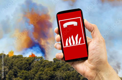 View on hand holding mobile smart phone with international fire emergency telephone symbol. Blurred fire flames background.