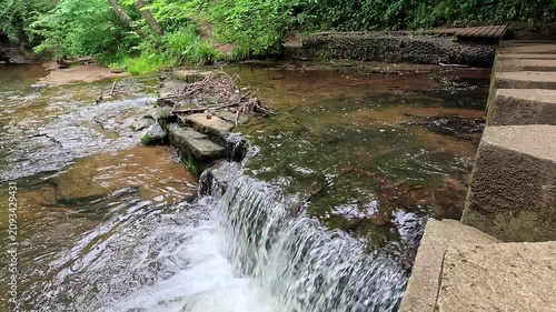 Stepping stones cross a woodland waterfall as the camera slowly pulls back in slow motion, revealing the flowing stream, mossy rocks, and tranquil forest surroundings in a serene natural scene.