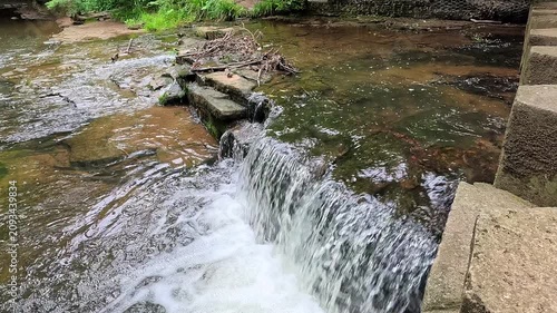 A slow-motion panning shot follows stepping stones across a woodland waterfall, capturing the flowing stream, mossy rocks, and lush forest surroundings in a peaceful natural landscape.
