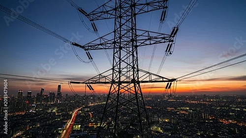 A large metal electricity pylon stands tall over a sprawling urban city at sunset