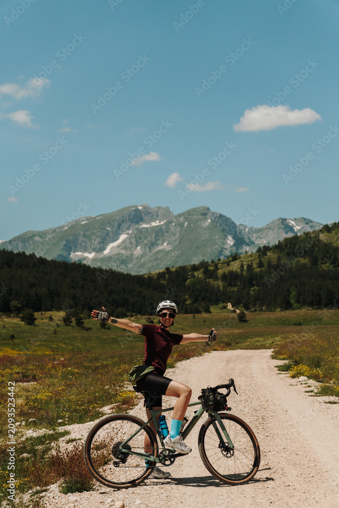 Fototapeta premium Happy woman posing with outstretched arms on a gravel bicycle in Durmitor National Park, Montenegro. Summer bikepacking adventure in alpine mountains.