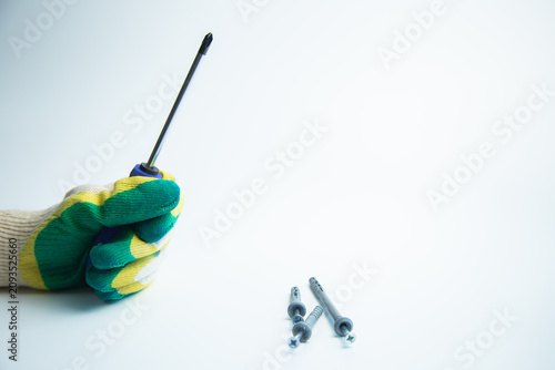A hand with a construction glove holds a screwdriver on a white background, next to it are self-tapping screws with space for text. Concept of repair, handyman, tools, construction, installation. High