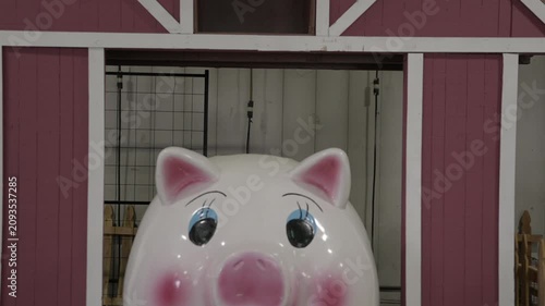 Upward tilt shot of a cute, oversized pink piggy bank sitting in the entrance of a traditional red rustic barn. 