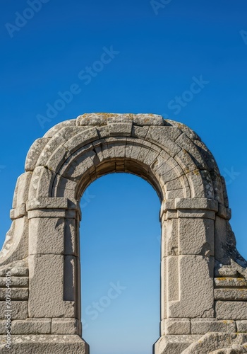 A massive, weathered stone archway stands resilient against a clear blue sky, showcasing timeless architectural strength and endurance, natural, tough, immemorial