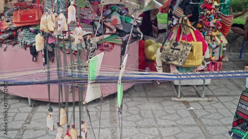 Close view of handloom threads under tension with hanging weights and subtle lateral shifting at outdoor market textile display beside colorful woven fabrics, with weaving, loom, thread, warp detail