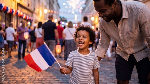 A laughing mixed-race toddler waves a French flag with his father during a joyful Bastille Day street festival for lifestyle stock photography