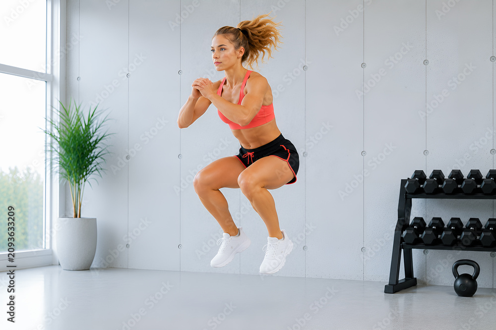 Fototapeta premium Kobieta podczas treningu wykonująca przysiad z wyskokiem w nowoczesnej domowej siłowni z hantlami i odważnikiem kettlebell.