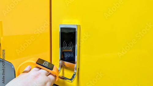 Fotografía hand bringing an orange RFID wristband to a contactless electronic lock on a bright yellow locker in a changing room