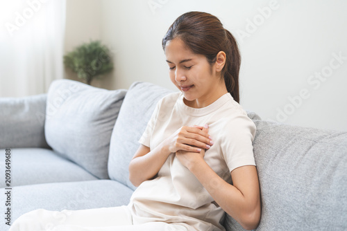 Obraz na plátně Heart attack, health care concept, suffering acute cardiac asian young woman hand touching on her chest pain, female breathing problem, asthma attack, pressure while sitting on sofa, couch at home