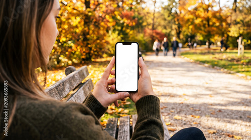 Hand holding smartphone with blank white screen on park bench, woman using mobile app for autumn stroll navigation, fall seasonal lifestyle, digital content mockup, nature path.
