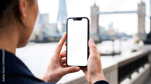 Hand holding smartphone with blank white screen, close up view in London, woman using mobile app for city travel guide mockup, urban tourism, digital navigation, river thames view.
