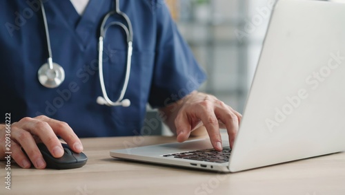 A close-up photo of a medical professional in blue scrubs with a stethoscope around their neck, typing on a silver laptop and using a black computer mouse on a wooden desk.
