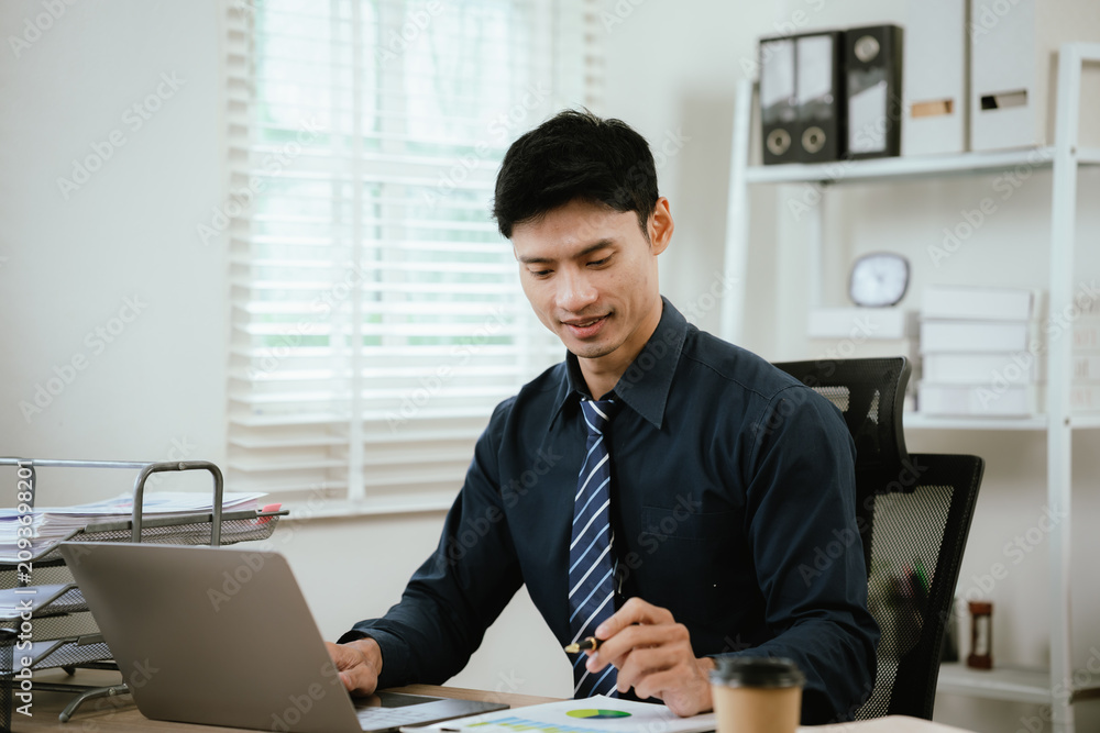 Fototapeta premium Young businessman working on a laptop in a modern office, reviewing documents and business reports during a productive workday.