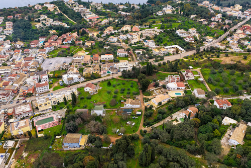 Panoramic aerial view of the coastal town of Kassiopi with the Ionian Sea in the background, surrounded by green hills and olive trees in Corfu, Greece.