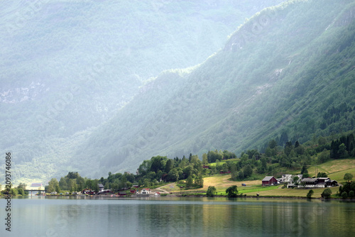 Sunbeams illuminate a peaceful fjord village in Norway, where lush mountains, calm water, and scattered farms create a serene Scandinavian landscape.