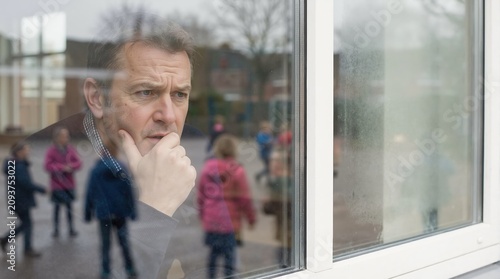 Serious man looking through window at school playground. Thoughtful male teacher or parent watching children play. Pensive man in educational environment