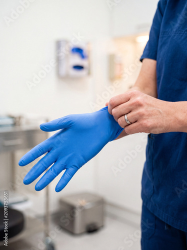 Healthcare professional putting on a blue medical glove in a clinical room. Close-up of hand hygiene and protective equipment preparation for safe patient care and medical procedures.