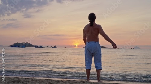 A man is warming up on the seashore at sunrise. A lone man stands on the shoreline facing the sea, silhouetted in warm sunset light. Calm waves, distant boats, and a small island create