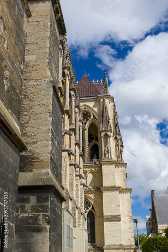 A side view of Reims Cathedral reveals ornate Gothic details rising sharply against a vivid blue sky. Stone buttresses and carvings create depth along the historic façade. Reims, France.