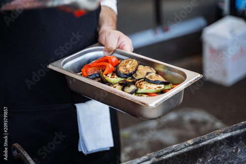 Cook holding a metal pan of grilled vegetables over a barbecue