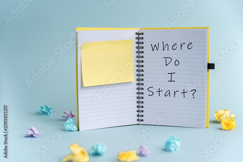 A spiral notebook with a yellow cover open to a page that reads 'Where Do I Start ' A sticky note is placed on the page. Colorful paper flowers surround the notebook.
