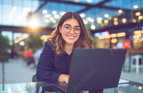 Smiling young woman with glasses working on a laptop at a modern cafe patio with glowing evening bokeh lights in the background