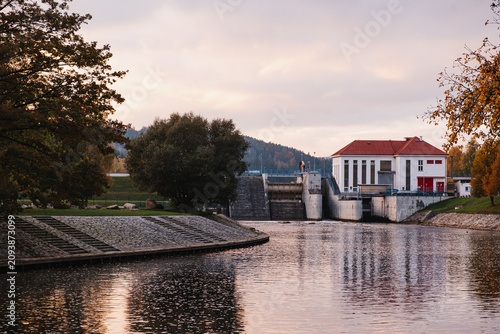 Hydroelectric dam and water control building by river at sunset with autumn trees, calm water reflections and industrial infrastructure in rural landscape.
