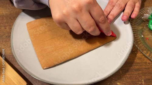 Horizontal 4K video close up. 60 fps. Top view. Woman draws red wave pattern on gingerbread house roof in plate on table. Female hand with plastic bag with edible paint. New Year 2027. Sweet food