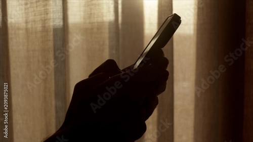 Silhouette of hand typing on smart phone near bright curtains