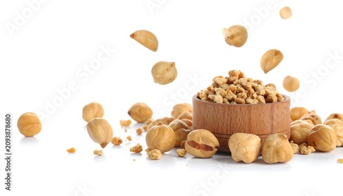 A wooden bowl filled with hazelnuts, some scattered around it on a white background