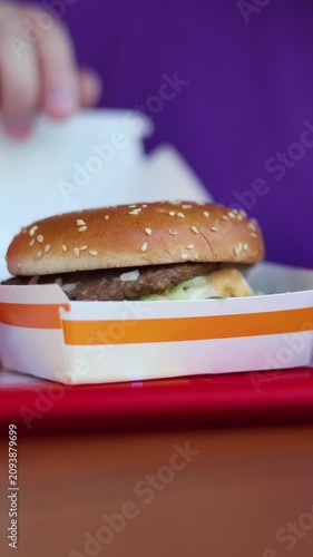 Vertical video close up. 60 fps. Fat man with blue t-shirt sits at fast food cafe table with French fries opens paper box with burger. Male hand touch burger in box. Concept of lunch, dinner, fastfood