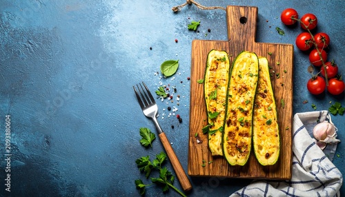 A wooden cutting board holds stuffed zucchinis, cherry tomatoes, and herbs on a blue surface