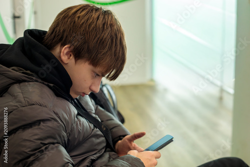 Konstfotografi Teenage boy in dark puffer jacket absorbed in smartphone while sitting in waitin