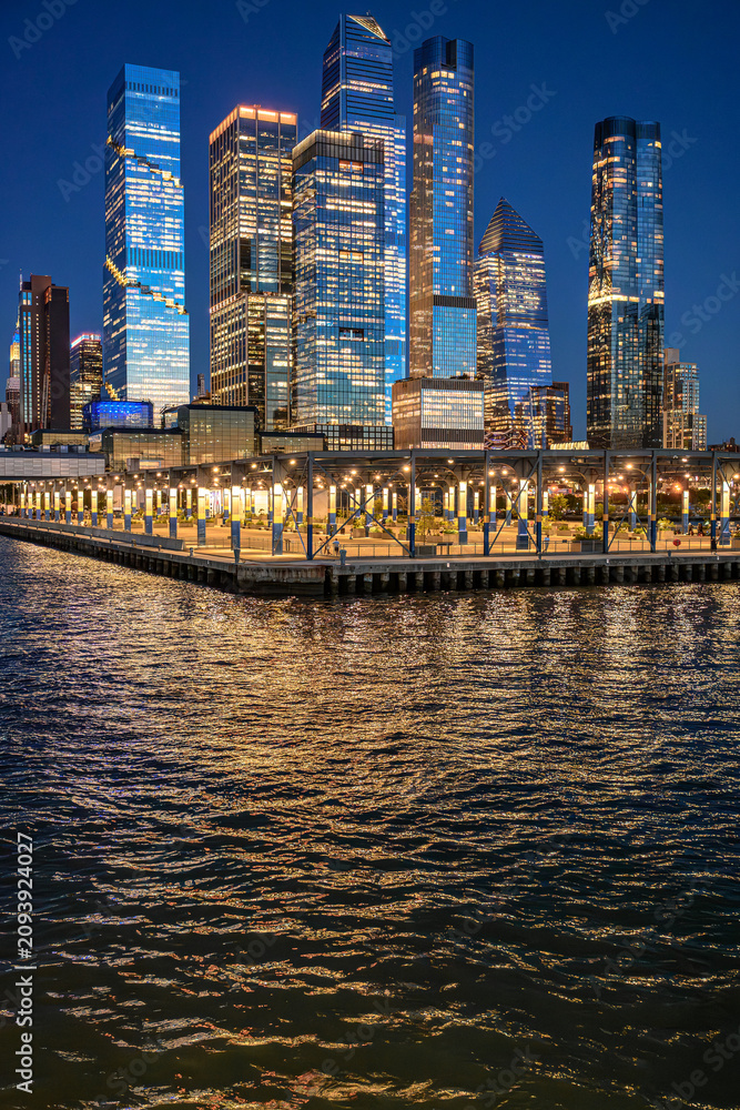 Fototapeta premium Hudson Yards skyline above Hudson River at blue hour. Illuminated Manhattan towers rise behind a covered waterfront pier with reflections on rippled water.