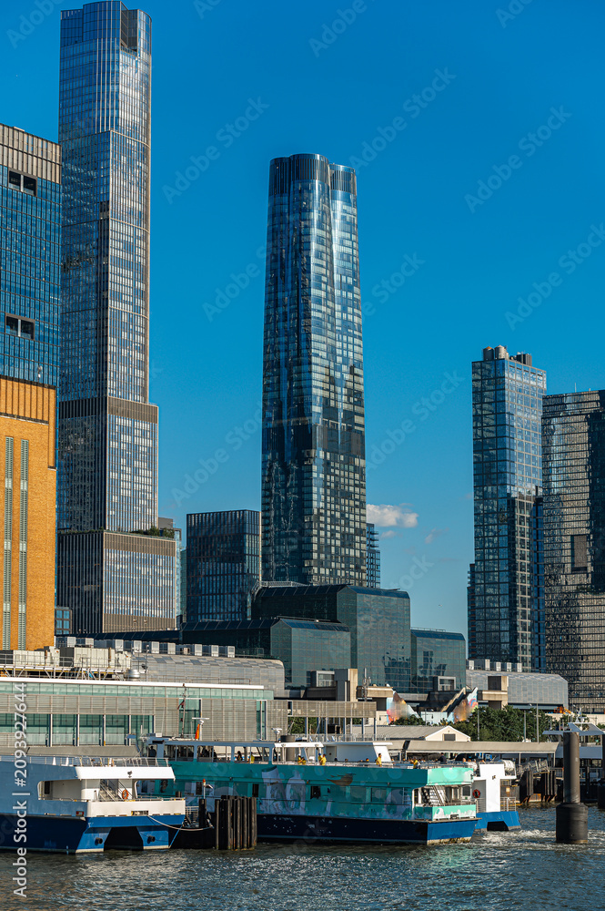 Fototapeta premium Passenger ferries below Hudson Yards residential towers, Manhattan. Vertical view frames glass high rises, terminal structures, dock pilings, and vessels along the river.