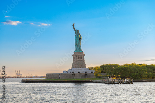 Statue of Liberty across New York Harbor at sunset. Front view shows the monument on Liberty Island with trees, docks, water, and port cranes behind.