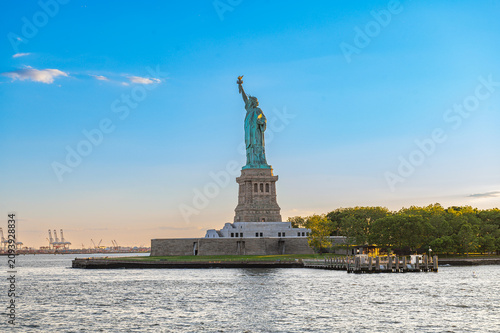 Statue of Liberty and Liberty Island in evening light. Copper monument rises above the stone pedestal, waterfront trees, ferry docks, and distant cranes.