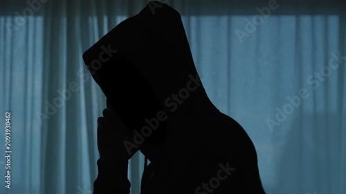 Hooded man silhouette making a suspicious phone call. Mysterious figure doing illegal activities blackmail or anonymous threats by backlit curtained window. Spy or hacker talking on mobile at night